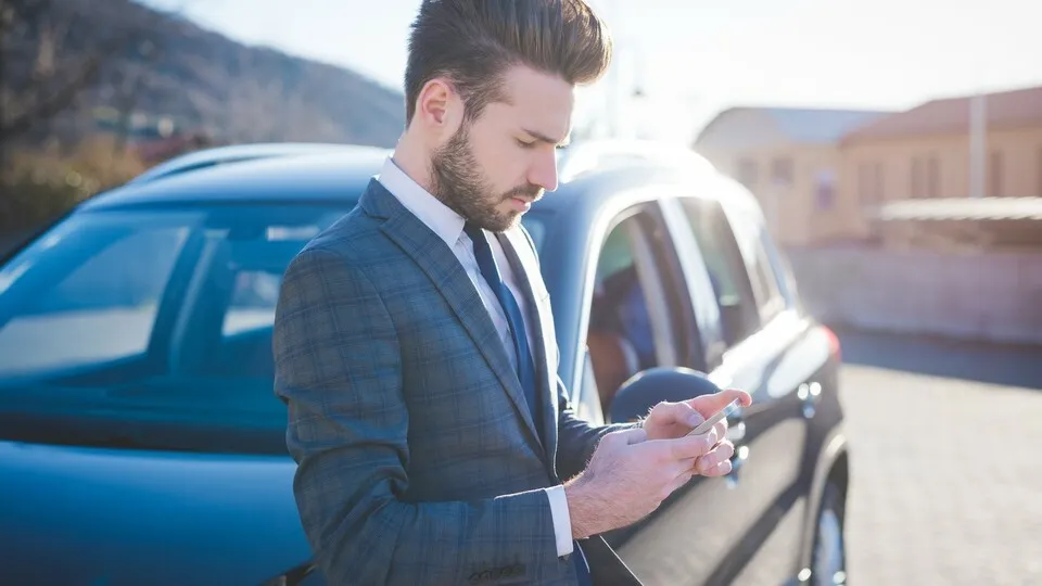05 Mar 2015 --- Young businessman leaning against car reading smartphone texts --- Image by © Eugenio Marongiu/Corbis